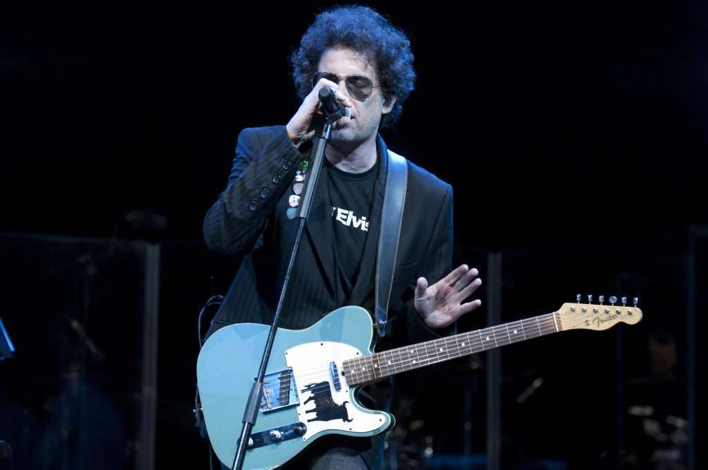 Andrés Calamaro cantando y tocando la guitarra en un concierto, con gafas de sol y una chaqueta negra.