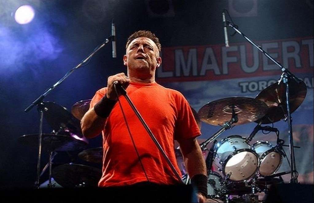 Ricardo Iorio en un concierto, sosteniendo un micrófono, con una batería en el fondo y luces de escenario.