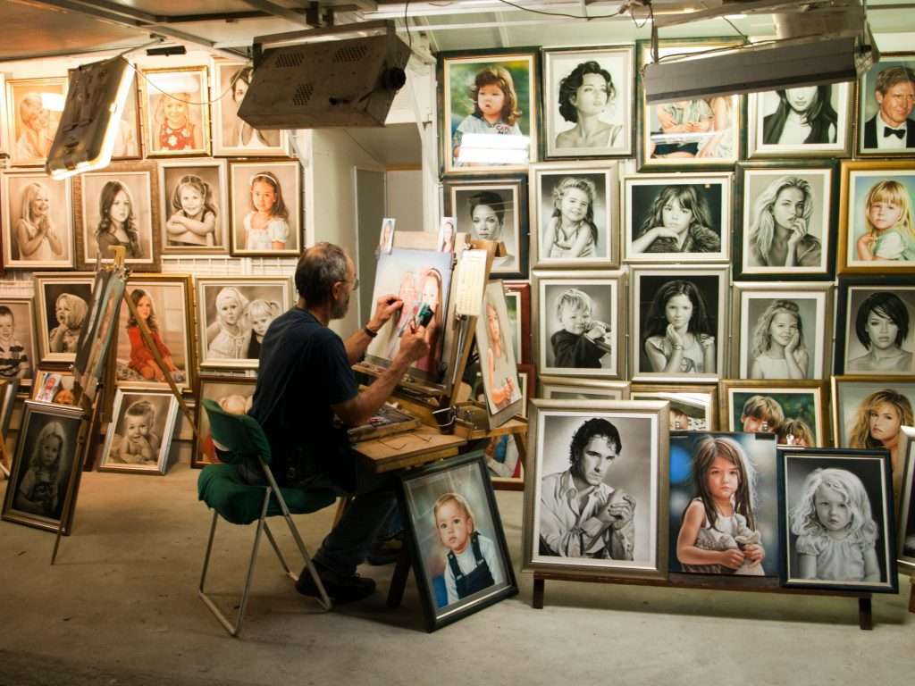 Artista trabajando en su estudio rodeado de retratos enmarcados de diversas personas, capturando diferentes expresiones y emociones.