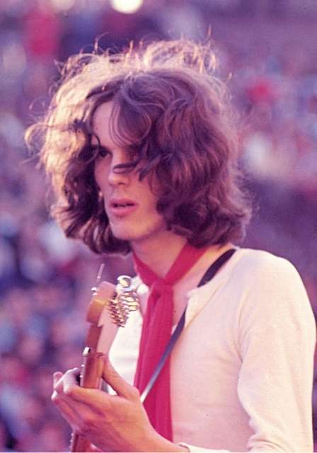 Luis Alberto Spinetta en un concierto al aire libre en 1969, sosteniendo una guitarra y usando una bufanda roja.